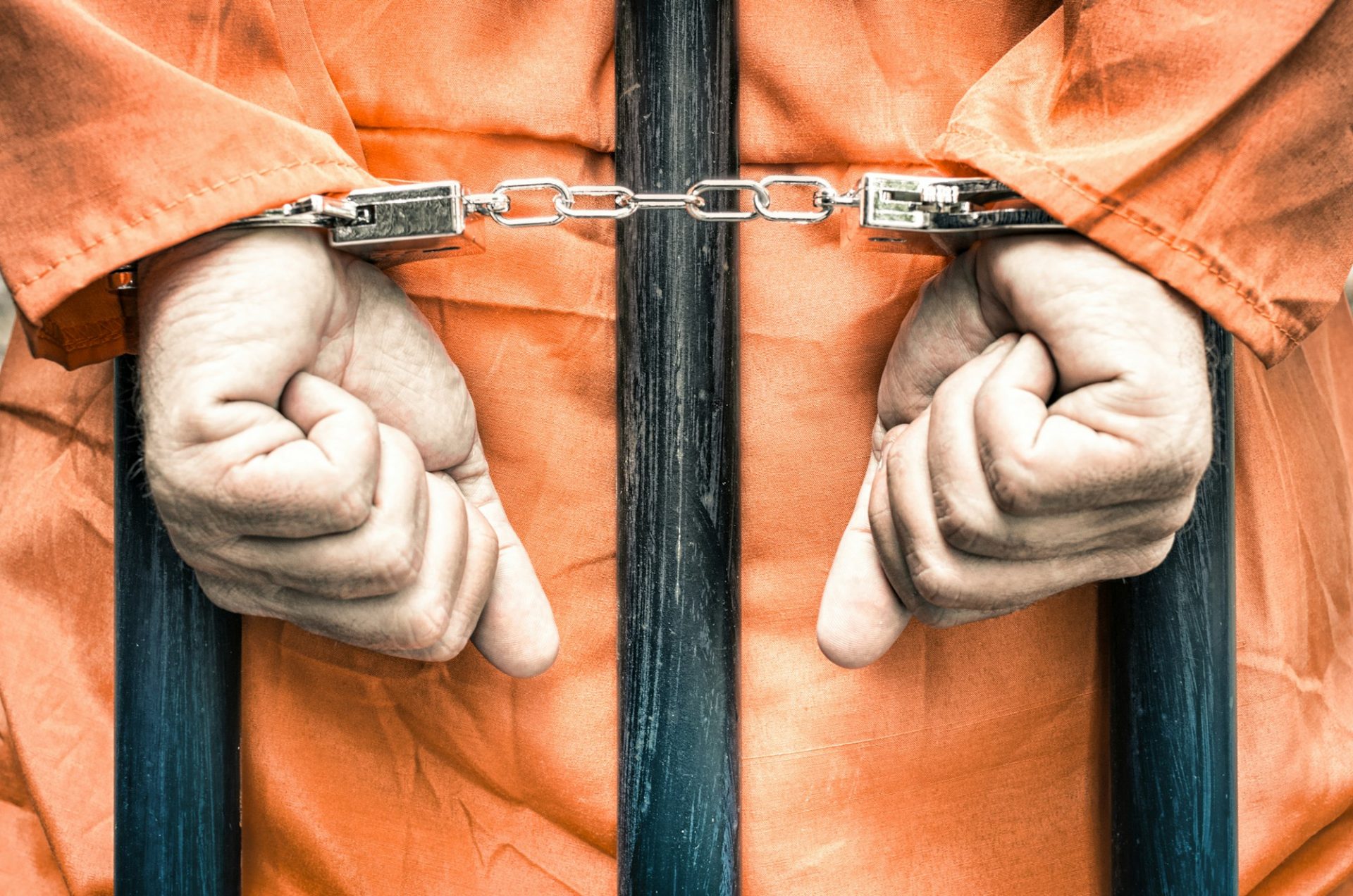 Handcuffed hands of a prisoner behind the bars of a prison with orange clothes